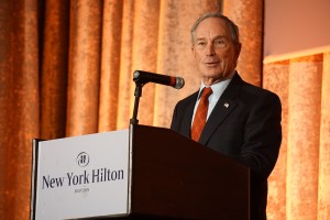 Michael Bloomberg stands behind a podium with a microphone and the New York Hilton logo in front of an orange backdrop